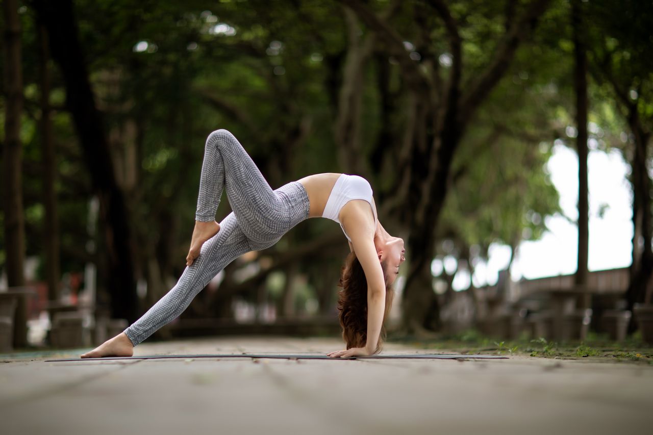 A woman performs a yoga pose outdoors, demonstrating flexibility on a tree-lined walkway.