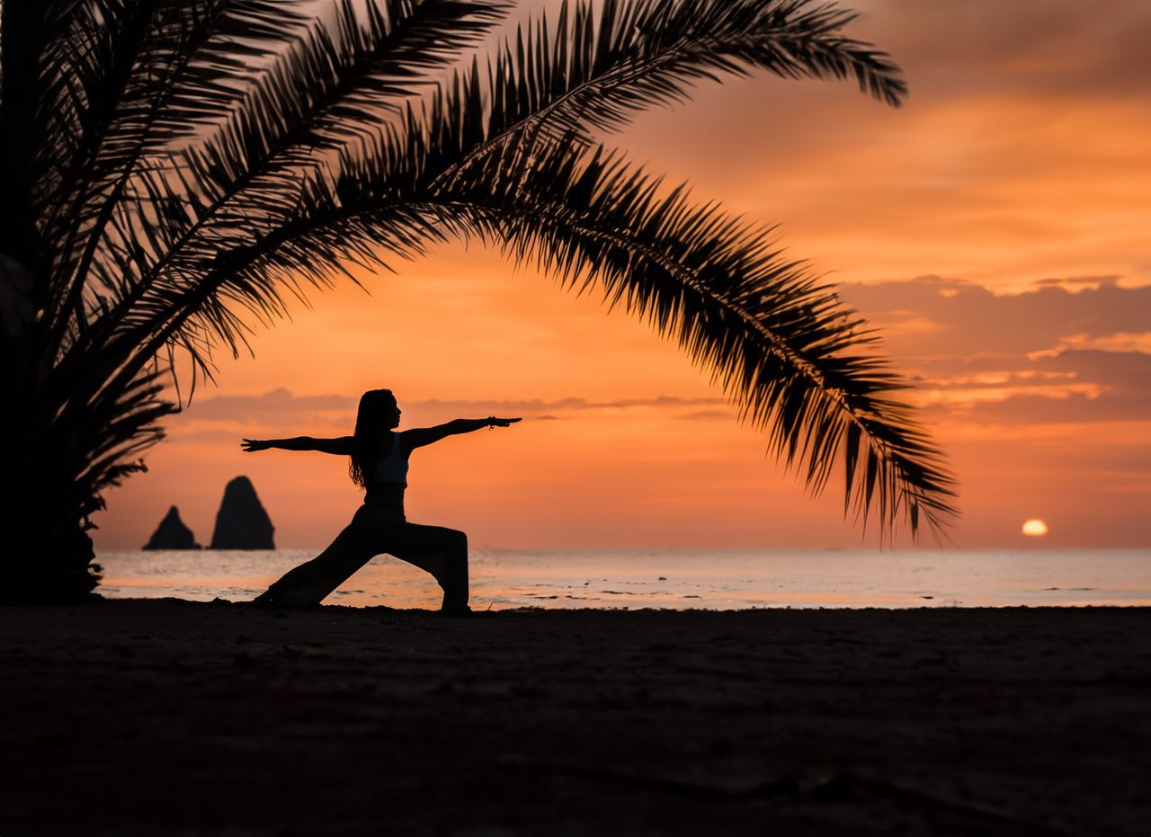 services-01 A woman practices yoga under palm trees during a vibrant sunset on a tropical beach.
